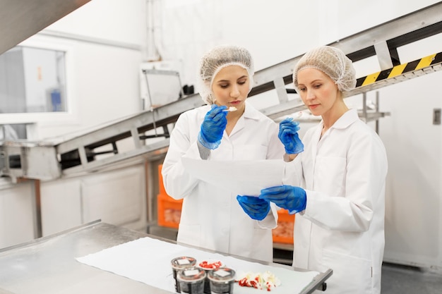 voedselproductie, industrie en mensenconcept - vrouwelijke technologen met een klembord proeven van ijs kwaliteit in de fabriek