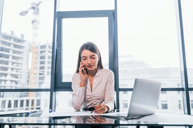 Voer een gesprek via de telefoon Jonge vrouw in witte formele kleding is binnenshuis in het moderne kantoor