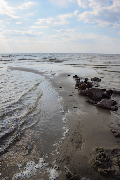 Voetafdrukken op nat zand bij een stapel stenen aan de kust met een branding tegen een bewolkte hemel