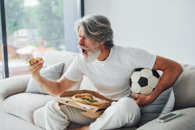 Voetbalfan kijkt naar wedstrijd en eet pizza Senior stijlvolle moderne man met grijs haar en baard binnenshuis