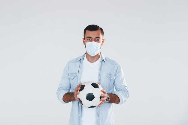 Voetbalfan met voetbal Jonge knappe man die binnenshuis tegen een witte achtergrond staat