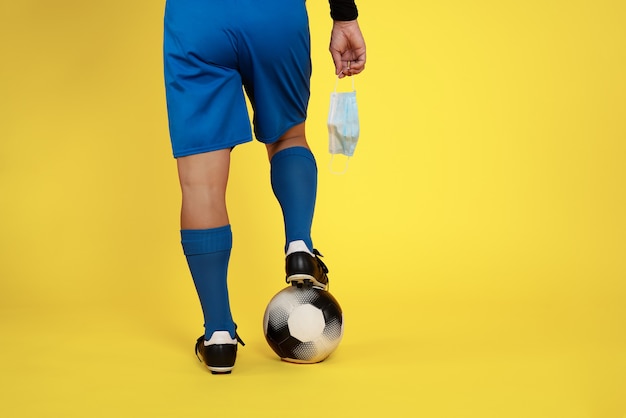 Foto voetballer met gezichtsmasker in de hand vanwege covid19 coronavirus pandemie en bal in voet op gele muur