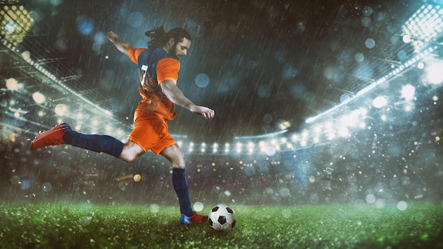 Voetbalscène bij nachtwedstrijd met speler in een oranje uniform die de bal met kracht schopt