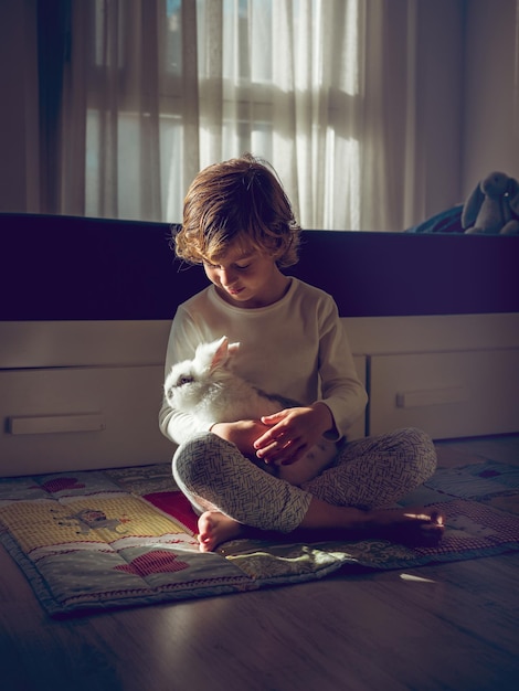 Vol lichaam van een schattig blootsvoetig kind in slaapkleding zit met een wit gedomesticeerd konijn in de armen bij het bed in een lichte slaapkamer