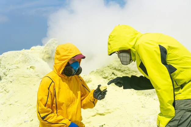 火山の斜面にいる火山学者は、硫黄の噴気孔を背景に鉱物のサンプルを研究しています