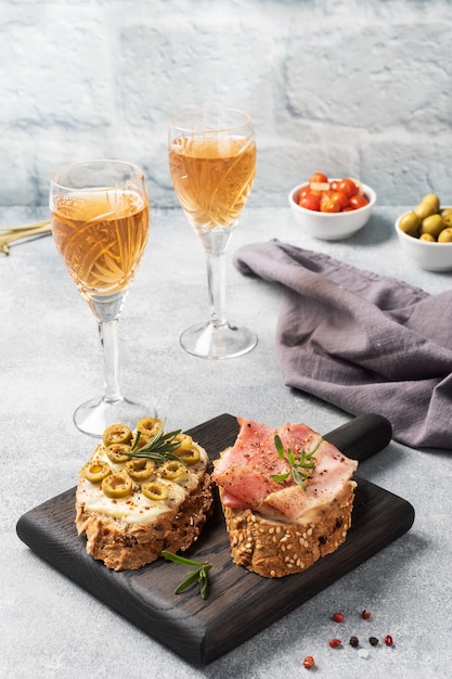 Volkoren brood sandwiches met roomkaas, spek en olijven op een houten snijplank. een glas witte wijn. grijze betonnen tafel met kopie ruimte.