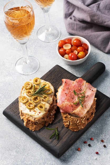 Volkoren brood sandwiches met roomkaas, spek en olijven op een houten snijplank. Een glas witte wijn. Grijze betonnen tafel.