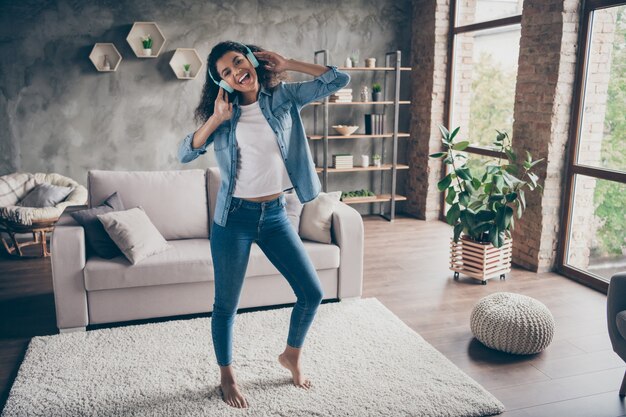 Volle lengte foto van mooie donkere huid golvende dame dragen moderne oorkleppen zingen luisteren coole liedjes staande vloer in de buurt van bank casual denim outfit woonkamer binnenshuis