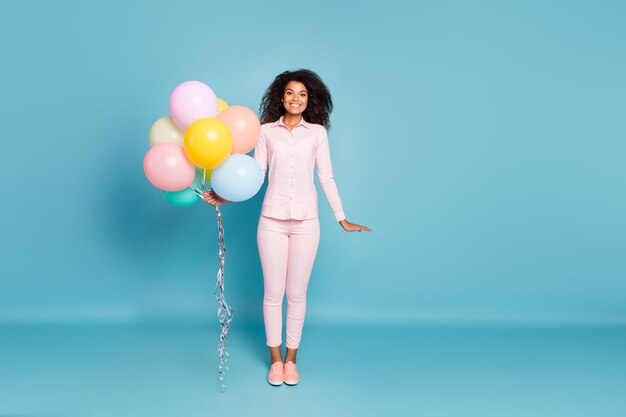 Volle lengte foto van verbazingwekkende golvende donkere huid dame met veel luchtballonnen opgewonden positieve feeststemming draag roze shirt broek geïsoleerde blauwe kleur achtergrond