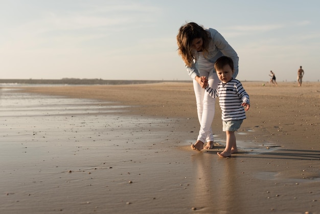 Foto volle lengte moeder helpt baby jongen lopen op het strand tegen de lucht
