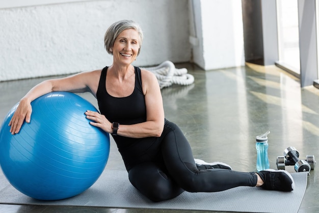 volle lengte van gelukkige senior vrouw in zwarte leggings en tank top zit op fitness m