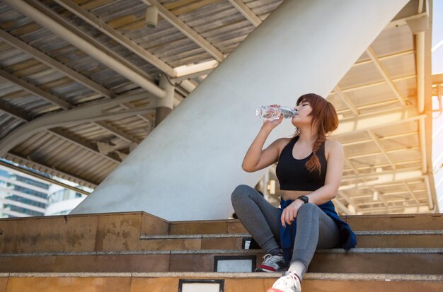 Foto volle lengte van jonge vrouw die water uit een fles drinkt terwijl ze op de trap zit