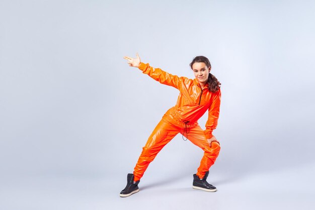 Volledig beeld van een krachtig tienermeisje met donkerbruin haar in een feloranje jumpsuit die hiphop danst en naar de zijkant wijst met een vingerpistool, hobbyactiviteiten. studio opname, witte achtergrond