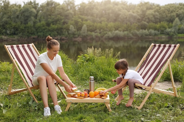 Volledig lengteportret van aantrekkelijke vrouw die wit t-shirt draagt dat op vouwende picknickstoelen met haar dochtertje zit en bananen, perziken, sinaasappelen en sandwiches eet