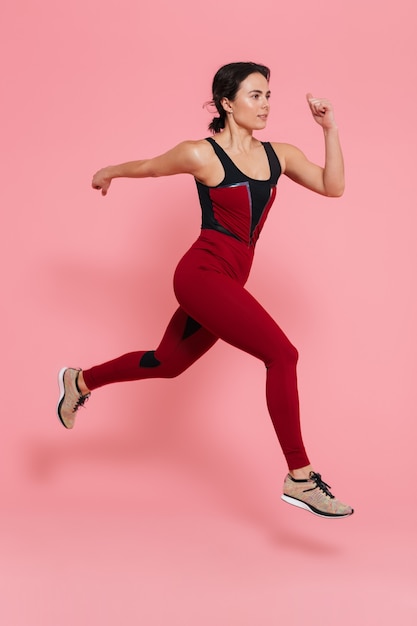 Volledig lengteportret van een geschikte mooie sportenvrouw die oefeningen doet die over roze muur worden geïsoleerd, springend