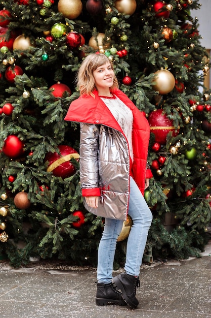 Volledig lengteportret van een jonge vrouw gekleed in een zilver en rood donsjack voor een feestelijk versierde kerstboom. Toeristische vrouw op kerstmarkt.