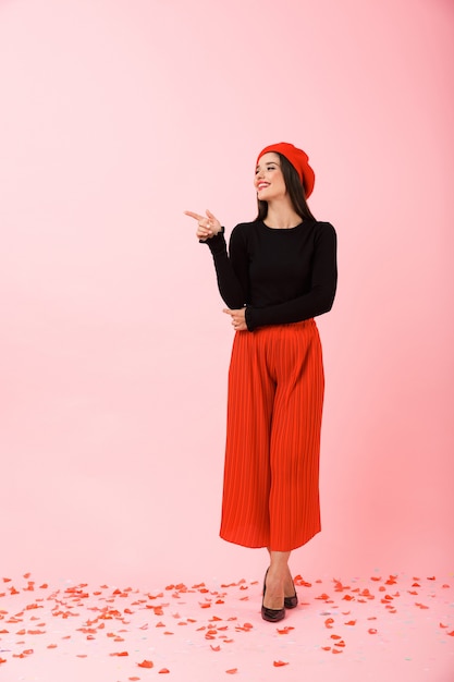 Volledig lengteportret van een mooie jonge vrouw die rode baret draagt die zich geïsoleerd over roze achtergrond bevindt, die exemplaarruimte voorstelt