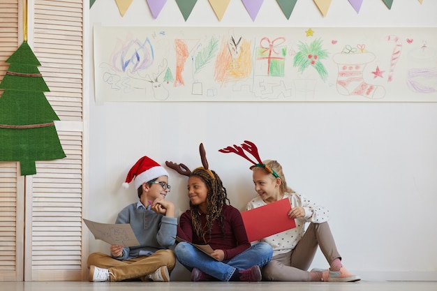 Volledig lengteportret van een multi-etnische groep kinderen die op vloer zitten die foto's houden terwijl u geniet van kunstles op Kerstmis, exemplaarruimte