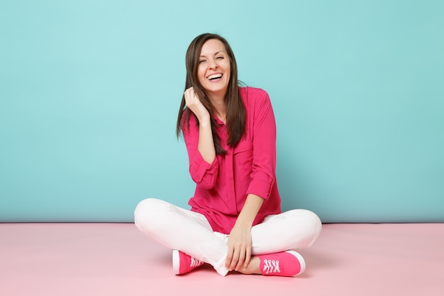 Volledig lengteportret van glimlachende jonge vrouw in roze overhemdblouse, witte broek die op vloer zit