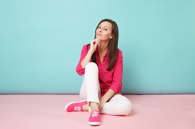 Volledig lengteportret van glimlachende jonge vrouw in roze overhemdblouse, witte broek die op vloer zit