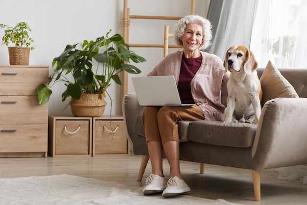 Volledig lengteportret van moderne hogere vrouw die laptop met behulp van terwijl zittend met hond op bank in gezellig appartement en glimlachen