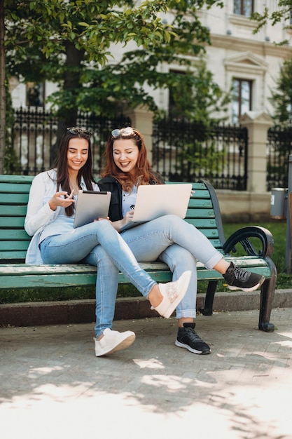 Volledig lengteportret van twee mooie vrouwenzitting op bank buiten die aan een tablet en laptop lacht en koffie drinkt.