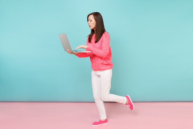 Volledig lengteportret van vrouw in gebreide roze trui, witte broek met behulp van laptop pc-computer