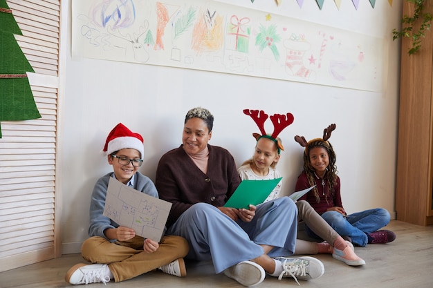 Volledig lengteportret van vrouwelijke leraar zittend op de vloer met een multi-etnische groep kinderen die foto's houden terwijl ze genieten van kunstles op Kerstmis, kopieer ruimte