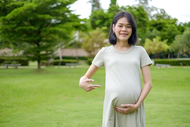 Volledig lichaam staat een zwangere moeder in een lange blauwe jurk en wijst met haar vinger naar haar buik in de tuin