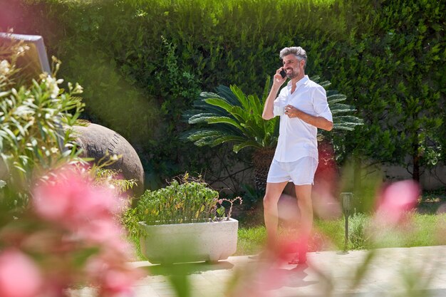 Volledig lichaam van bebaarde man in vrijetijdskleding die in de buurt van groene planten in de tuin loopt en op smartphone spreekt