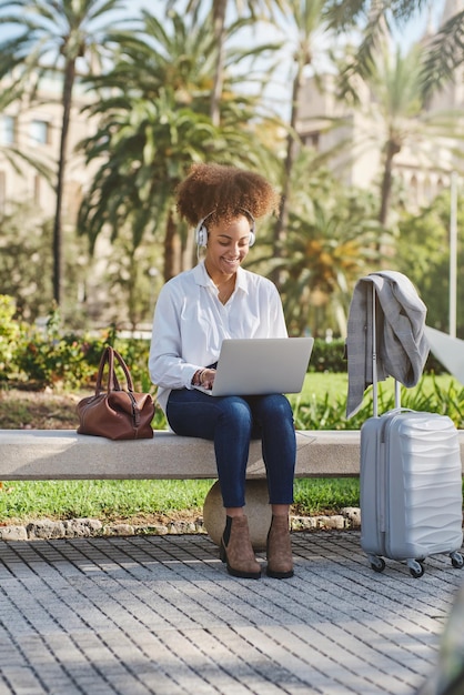 Volledig lichaam van glimlachende Afro-Amerikaanse vrouw in koptelefoon zittend op een bankje en luisterend naar muziek terwijl ze op afstand werkt aan netbook in groen stadspark