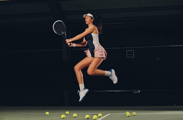 Volledig lichaamsportret van vrouwelijke tennisspeler in een sprong op een tennisbaan over donkere achtergrond.