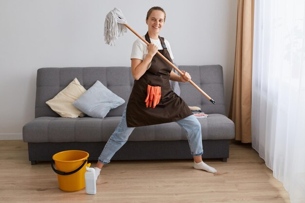 Volledig portret van een aantrekkelijke, gelukkig lachende vrouw met een spijkerbroek en een bruin schort met een dweil in handen en dansend tijdens het wassen van de vloer in de woonkamer