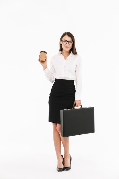 Volledig portret van een aantrekkelijke jonge zakenvrouw in formele kleding die over een witte muur staat, aktetas draagt, afhaalkoffie drinkt