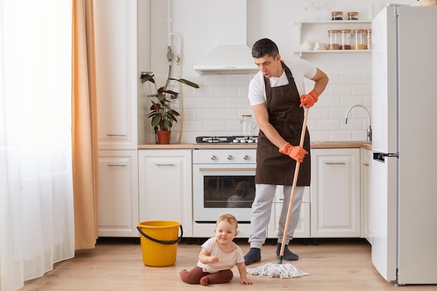 Volledig portret van een aantrekkelijke, vrolijke man die de vloer schoonmaakt in het huis dat de keuken schoonmaakt en voor peuterdochter zorgt die zijn vrouw helpt met huishoudelijke taken