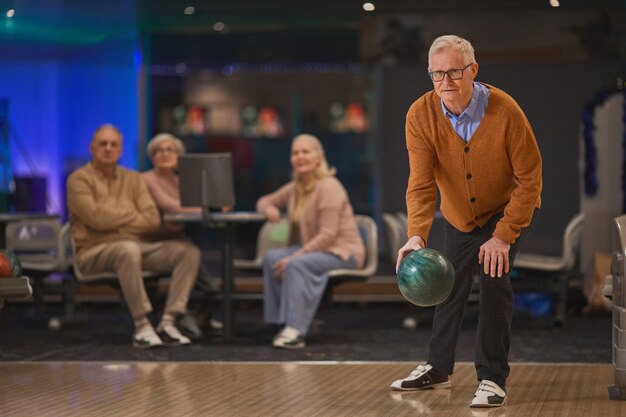 Volledig portret van een actieve senior man die bowlen speelt met een groep vrienden op de achtergrond, kopieer ruimte