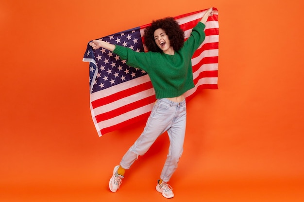 Volledig portret van een gelukkige vrouw met Afro-kapsel in een groene, casual stijl die doet alsof ze vliegt, plezier heeft en een Amerikaanse vlag vasthoudt. Indoor studio opname geïsoleerd op een oranje achtergrond.