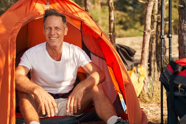 Volledig portret van een glimlachende sportieve man die op de camping zit na nordic walking in de natuur