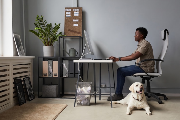 Foto volledig portret van een jonge afro-amerikaanse man die op kantoor werkt met een hond die op de vloer ligt, huisdiervriendelijke werkruimte, kopieerruimte