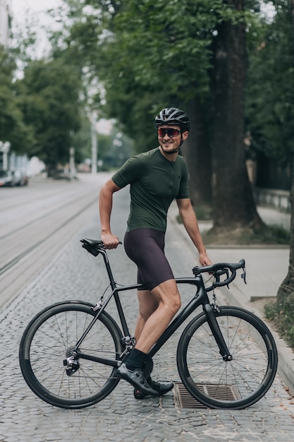 Volledig portret van een jonge man in sportkleding, helm en bril die opzij kijkt terwijl hij op de fiets rust na een training in de buitenlucht. Concept van mensen, actieve hobby en recreatie.