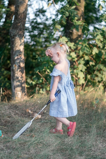 Volledig portret van een klein meisje in blauwe zomerjurk met racket in haar handen Kind speelt buiten badminton