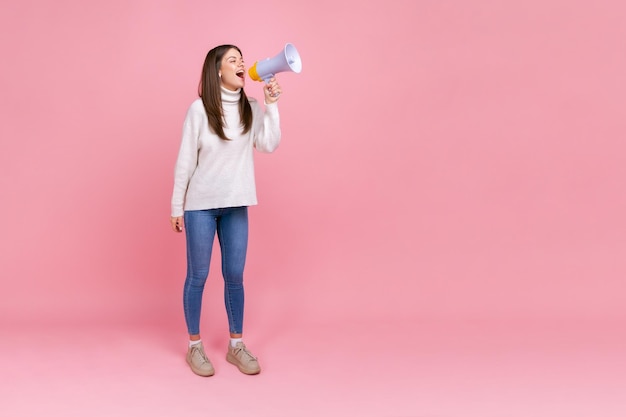 Volledig portret van een mooie vrouw die luid schreeuwt in een megafoon, belangrijke informatie aankondigt, met een witte casual stijltrui. Indoor studio opname geïsoleerd op roze achtergrond.