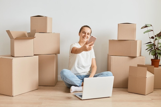 Volledig portret van een opgetogen vrouw met een spijkerbroek en een wit t-shirt die op de vloer in de buurt van kartonnen dozen zit en een videogesprek heeft of livestream uitzendt terwijl ze naar een nieuw appartement verhuist