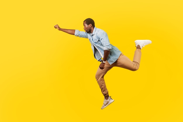 Volledig portret van een serieuze, coole knappe man in een denim casual shirt dat in de lucht springt of hoog vliegt met een gestrekte arm, het gevoel superman te zijn. indoor studio-opname geïsoleerd op gele achtergrond