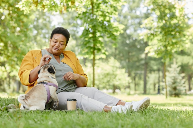 Volledig portret van een zwarte oudere vrouw die met een schattige hond in het park speelt terwijl ze op het gras zit en