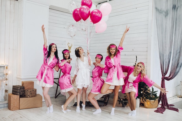 Volledig portret van gelukkige mooie vriendinnen in roze gewaden en slaapmaskers die springen en plezier hebben op vrijgezellenfeest bridetobe in wit gewaad met roze luchtballonnen met een glimlach die op hun tenen staat