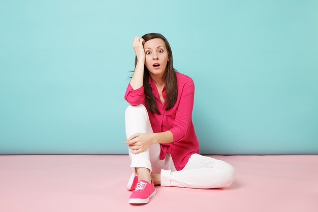 Volledig portret van geschokte jonge vrouw in roze overhemdblouse, witte broek zittend op de vloer