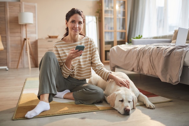 Volledig portret van glimlachende jonge vrouw zittend op de vloer met smartphone en aaiende hond in een gezellig interieur, kopieer ruimte