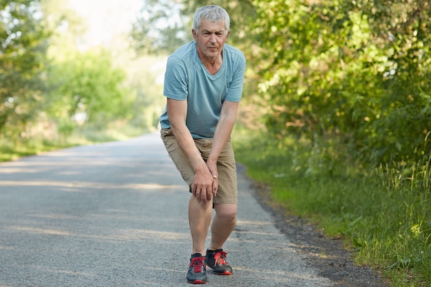 Volledig portret van ontevreden mannelijke gepensioneerde, leunt op knieën, heeft pijn na joggen, sloeg met zijn been, staat op de weg in landelijk gebied