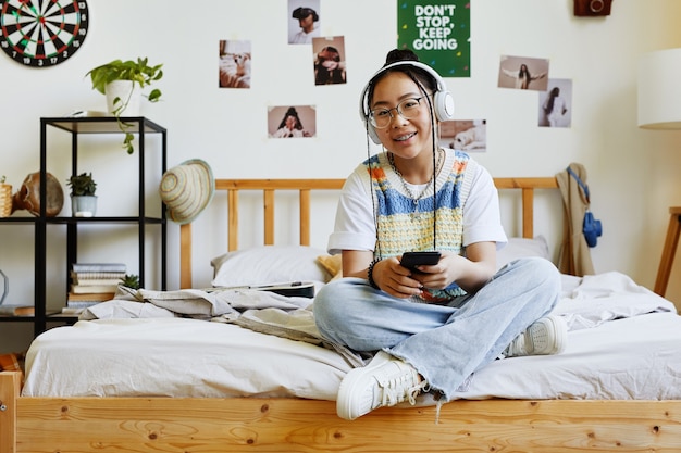 Volledig portret van trendy tienermeisje dat naar de camera kijkt terwijl ze op bed in een gezellige kamer zit, kopieer ruimte